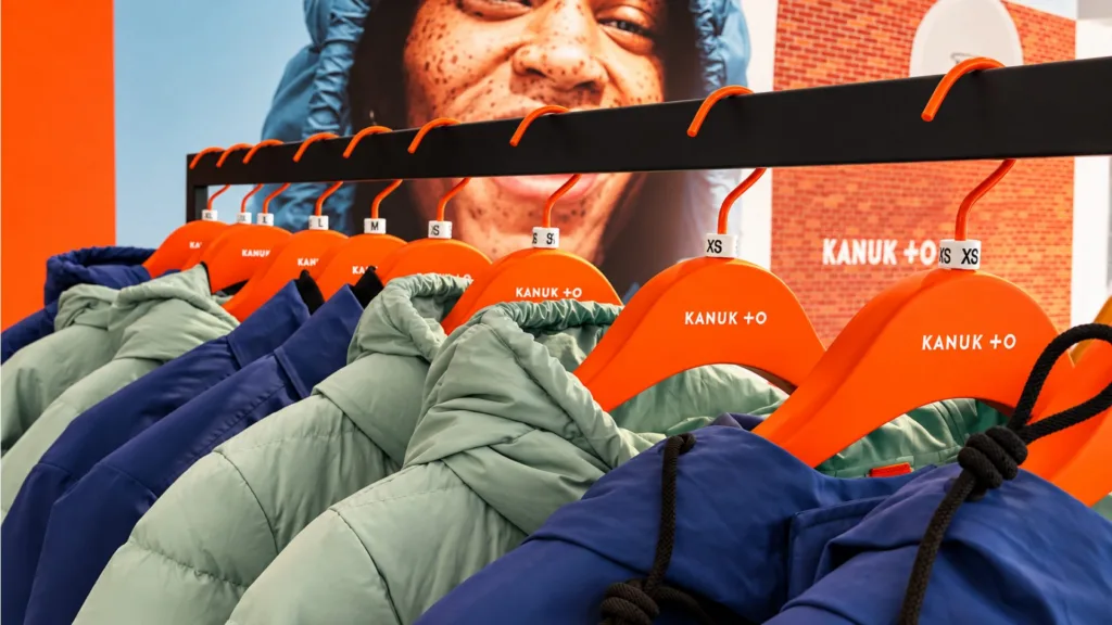 Manteaux KANUK + O sur cintres orange, devant une affiche